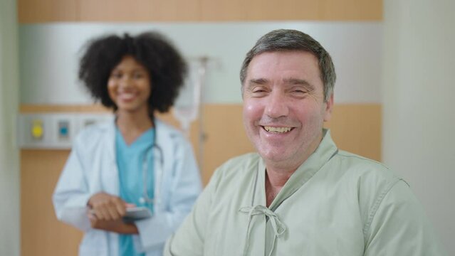 Smiling Patient Receiving Medical And Looking At Camera, African American Black Woman Doctor Standing Behind In Hospital Room. Elderly People Healthcare Concept.