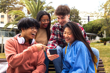 Happy young college student friends look at mobile phone. Multiracial teenagers using smartphone laugh together outdoor.