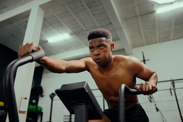 Plakat African American male using an elliptical trainer during cross fit training. Male athlete exercising intensely in the gym. High quality photo