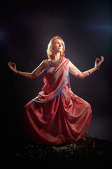 A beautiful adult skinny European blonde woman in a beautiful Indian sari does yoga exercises on a black background in the studio. Sporty romantic girl model posing in a photo shoot