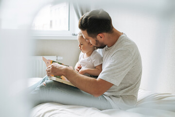 Happy father young man and baby girl little daughter having fun reading a book in children room at...