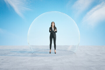Attractive happy european businesswoman with folded arms standing under and inside big glass sphere cover on concrete ground and bright blue sky background. Protection and limitation concept.
