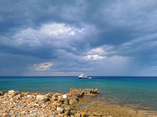 The stone coast of the Mediterranean Sea against the backdrop of the sea with a walking white ship and a dramatic sky.