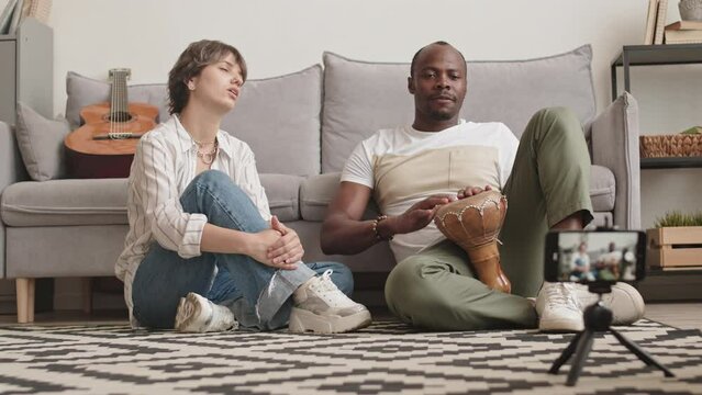 Stab shot of young diverse couple of bloggers recording video of themselves playing djembe and singing, sitting on rug in living room