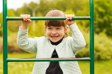 Charming 4-year-old Caucasian little girl having fun on the playground and smiling. A cheerful little girl is standing on the step of the stairs on the playground. Happy childhood.Active outdoor games