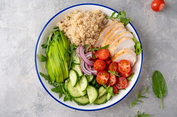 Healthy bowl lunch with grilled chicken, quinoa, avocado, tomatoes, cucumbers and fresh arugula on gray background. Top view. Selective focus.