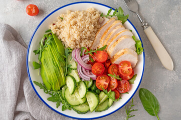 Healthy bowl lunch with grilled chicken, quinoa, avocado, tomatoes, cucumbers and fresh arugula on gray background. Top view. Selective focus.