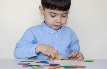 cute serious face little boy at white table playing with tangram puzzle.puzzle of seven flat polygons,tans,form shapes.educational,logic game for kids.back to school,kindergarten