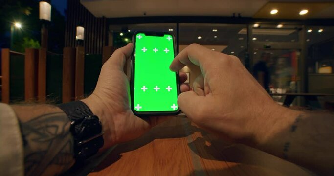 Close up front view of young male tattooed hands holding vertical and using mobile phone with green screen, swiping or scrolling smartphone touchscreen on table with soft lights of cafe on background