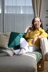 Obraz premium Smiling middle aged woman enjoying pleasant conversation and looking through the window while resting sofa at home