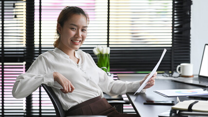 Confident young female manager sitting at her workplace, holding document and smiling a camera