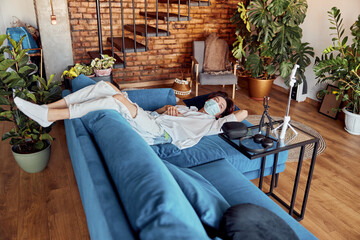 Guy wearing medical mask during virus epidemic lockdown and lying on sofa