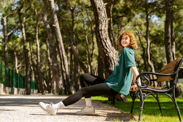 Naklejka na ściany i meble Happy redhead woman wearing green tee standing on city park, outdoor concentrating on her workout, leaning on a park bench, doing triceps exercise.