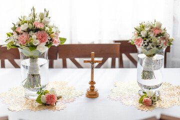 Fototapeta na wymiar Soft bridal bouquets of fresh flowers in glass vases and church cross on celebratory table in restaurant. Elegant attribute and tradition. Beautiful and romantic wedding, luxurious celebration