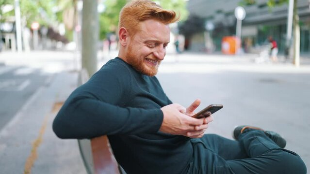 Smiling ginger man texting by smartphone while sitting on the bench outdoors
