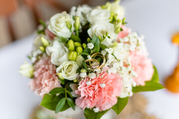 Soft bridal bouquet of fresh flowers and wedding gold rings on celebratory table closeup, blurred background. Elegant attribute and tradition. Beautiful and romantic wedding, luxurious celebration