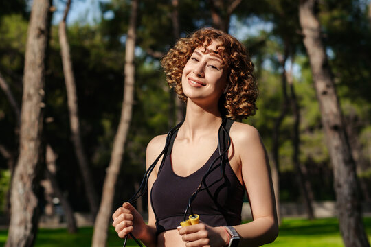 Portrait of cute sportive woman wearing sports bra standing on city park, outdoors holding a skipping rope on her neck looking at the camera during work out.