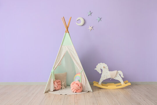 Children's wigwam with toys and rocking horse near violet wall in room