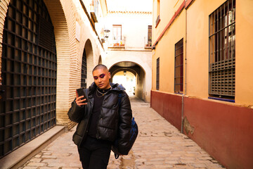 Fototapeta na wymiar Non-binary and young person from South America is on holiday in Europe, the person is make up and wearing black clothes. He is walking around the city consulting his mobile phone. Concept of equality.