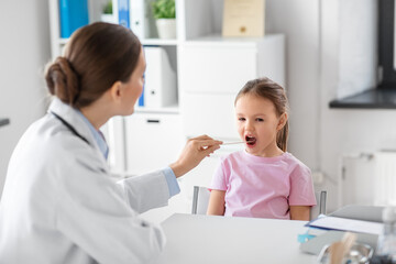 medicine, healthcare and pediatry concept - female doctor or pediatrician with tongue depressor checking little girl patient's throat on medical exam at clinic