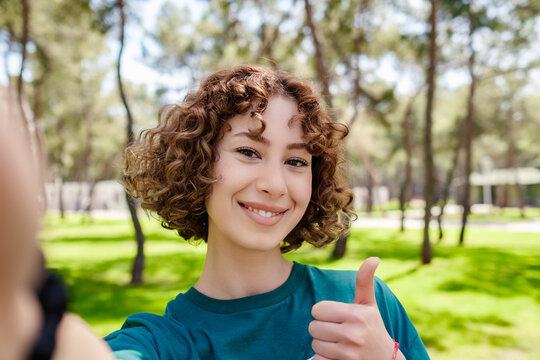 Happy redhead woman wearing tee standing on city park, outdoors taking a self portrait with smart phone. She looks at the screen and taking selfie. Thumbs up.