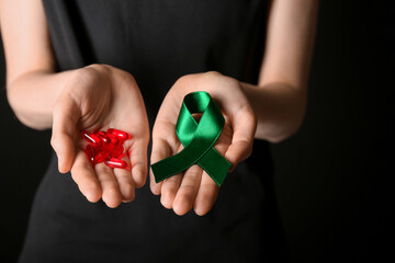 Woman with green ribbon and pills on black background, closeup. Liver cancer concept