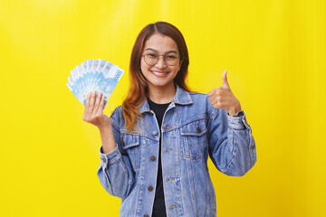 Smiling young asian woman standing while holding an Indonesian money and showing thumbs up