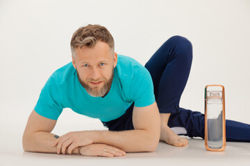 Young athletic man in blue sport suit lie down benting over near bottle of water closeup, white background. Keeping fit by fitness workout in gym, gymnastics. Sport and healthy lifestyle concept