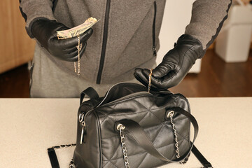 Robber in black outfit and gloves see in opened stolen women bag. The thief takes out the gold and...