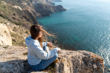 Fototapeta na wymiar Woman tourist enjoying the sunset over the sea mountain landscape. Sits outdoors on a rock above the sea. She is wearing jeans and a blue hoodie. Healthy lifestyle, harmony and meditation
