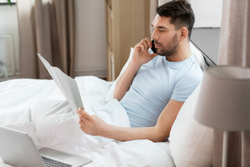 people, technology and remote job concept - man with folder and laptop computer calling on smartphone in bed at home bedroom