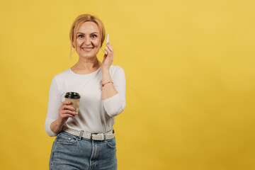 Attractive mature women over isolated yellow background holding coffee to take away and a mobile. Senior woman 50s. Copy space