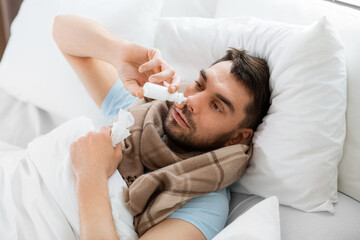 Obraz na płótnie Canvas people and health problem concept - unhappy sick man spraying his nose with nasal spray lying in bed at home