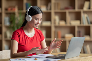 Smiling pretty teen chinese girl in headphones with laptop have online call and gestures in room interior