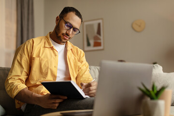 Focused young African American man using laptop, working online, taking notes during business meeting at home