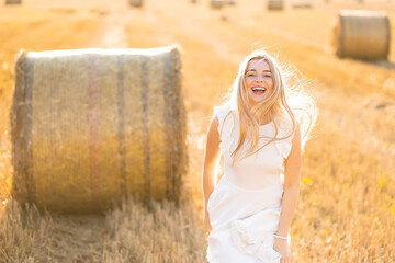 beautiful young woman with long hair in a white dress looking at the camera in a summer field near a haystack. Sunny day. rustic rural landscape © Ivan