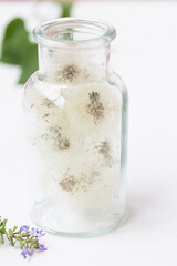 A glass bottle, vase with white blowballs, dandelions inside on the table, green leaves. Summer decoration