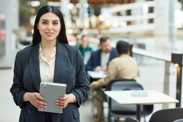 Waist up portrait of young Middle Eastern businesswoman holding planner and smiling at camera in modern office building interior, copy space
