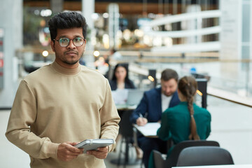 Waist up portrait of young man of Indian ethnicity holding planner and looking at camera in office building setting, copy space