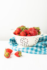 View of strawberries in a white colander on a white table with a blue kitchen cloth, white background with three strawberries, vertical