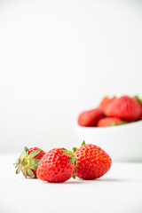 Close-up of three strawberries on a white table with a bowl in the background, selective focus, white background, vertical, with copy space