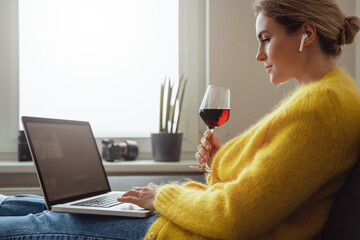 Woman sitting on the sofa drinking red whine while watching movie or working on her laptop at home