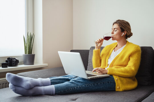 Woman Sitting On The Sofa Drinking Red Whine While Watching Movie Or Working On Her Laptop At Home