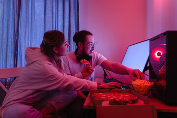 Couple chilling at home in room with neon light and eating pizza, watching movies online or playing video games