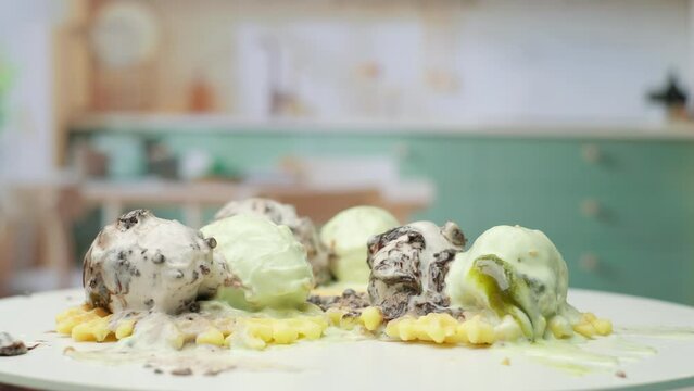 Melting pistachio and chocolate ice ream balls on the top of waffles rotating in the kitchen in slow motion.