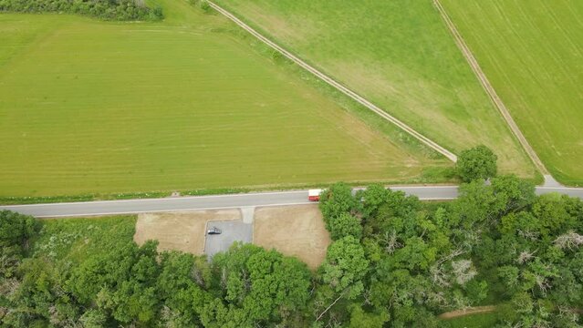 A red 5 ton truck driving along an empty curved road in between lush green meadows and thick leafy trees. Aerial parallel following top view