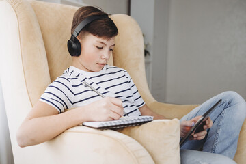 Obraz premium Teenage boy in wireless headphone using tablet studying online at home. Distance online education. Concept of e-learning, homeschool, virtual school.