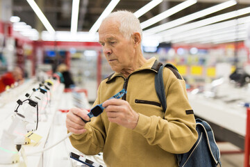 elderly grayhaired man pensioner examining counter with electronic gadgets and smart watches in...