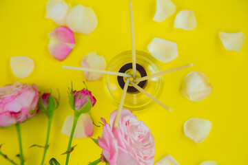 A glass jar with scented oil and wooden incense sticks in the room, next to roses and rose petals on a bright yellow background. High quality photo