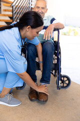 Biracial female physiotherapist adjusting legs of caucasian senior man sitting on wheelchair at home